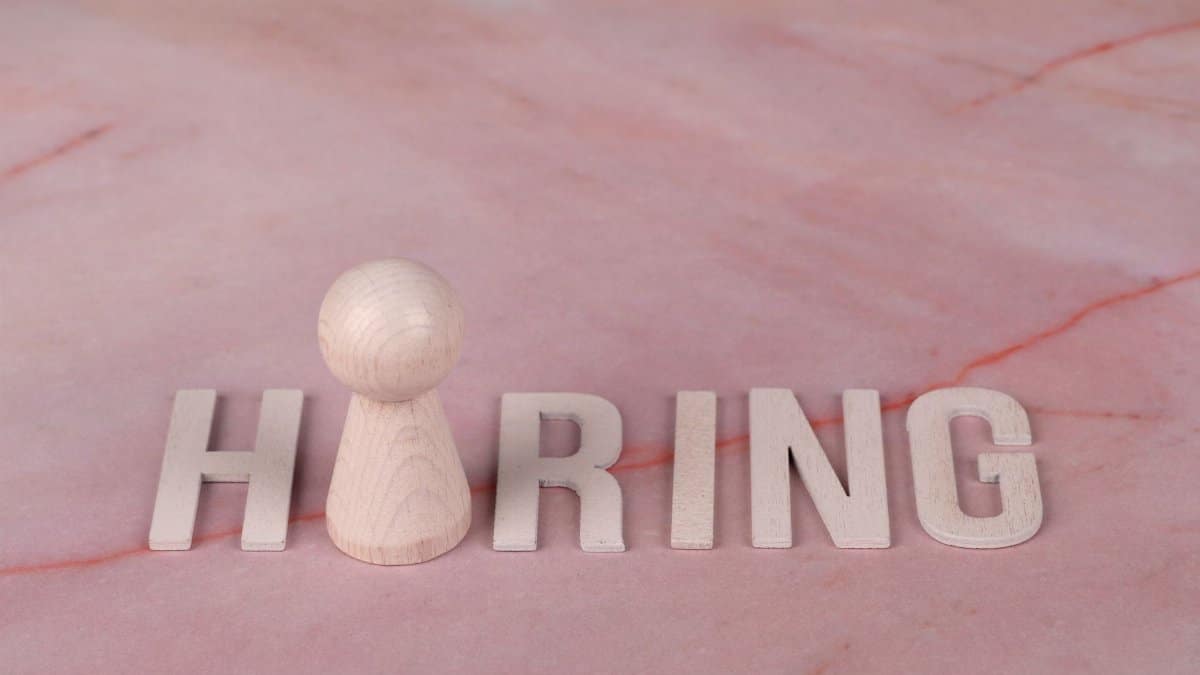 Wooden letters spelling 'hiring' on pink marble background with a peg.