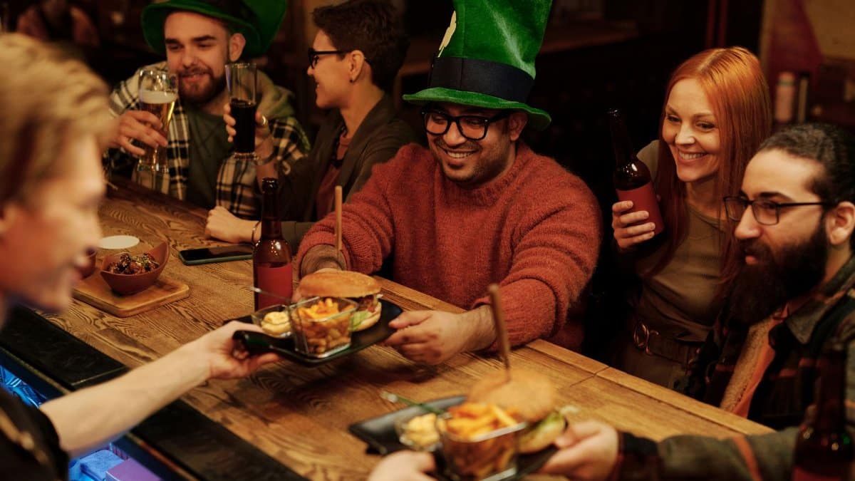 Friends wearing green hats enjoy food and drinks at a festive bar celebration.