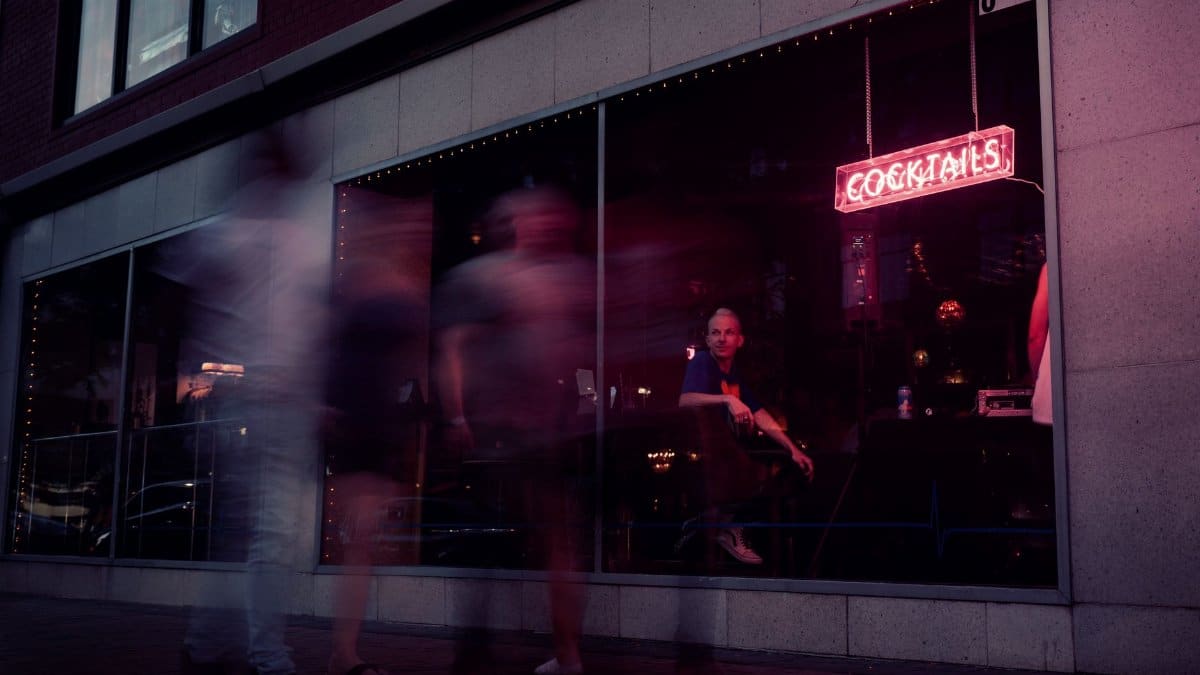 Blurred people walking past a bar with a neon cocktail sign at dusk.
