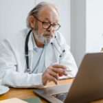 Senior male doctor using laptop for telehealth consultation in office setting.