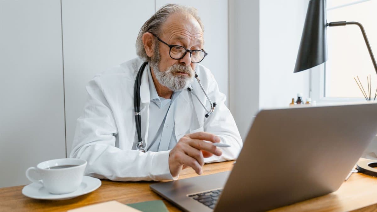 Senior male doctor using laptop for telehealth consultation in office setting.