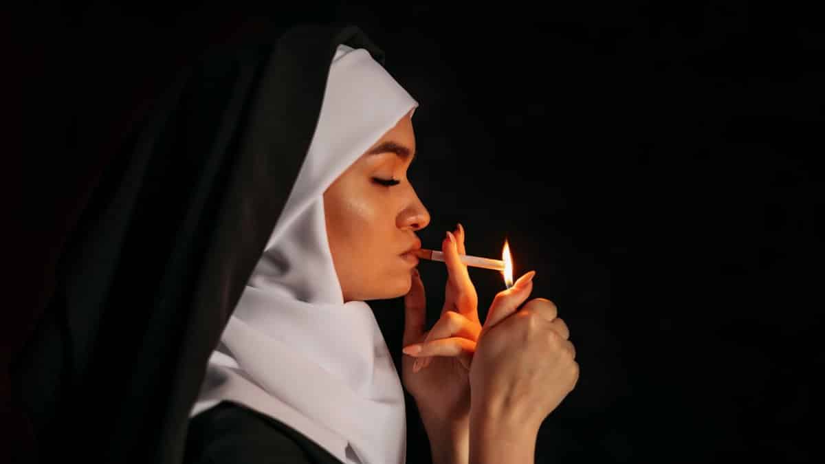 Side view portrait of a nun in a habit lighting a cigarette against a dark background, exploring contrasts.