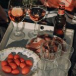 A beautifully arranged outdoor picnic with wine glasses, tomatoes, cold cuts, and a beer bottle on a tray.