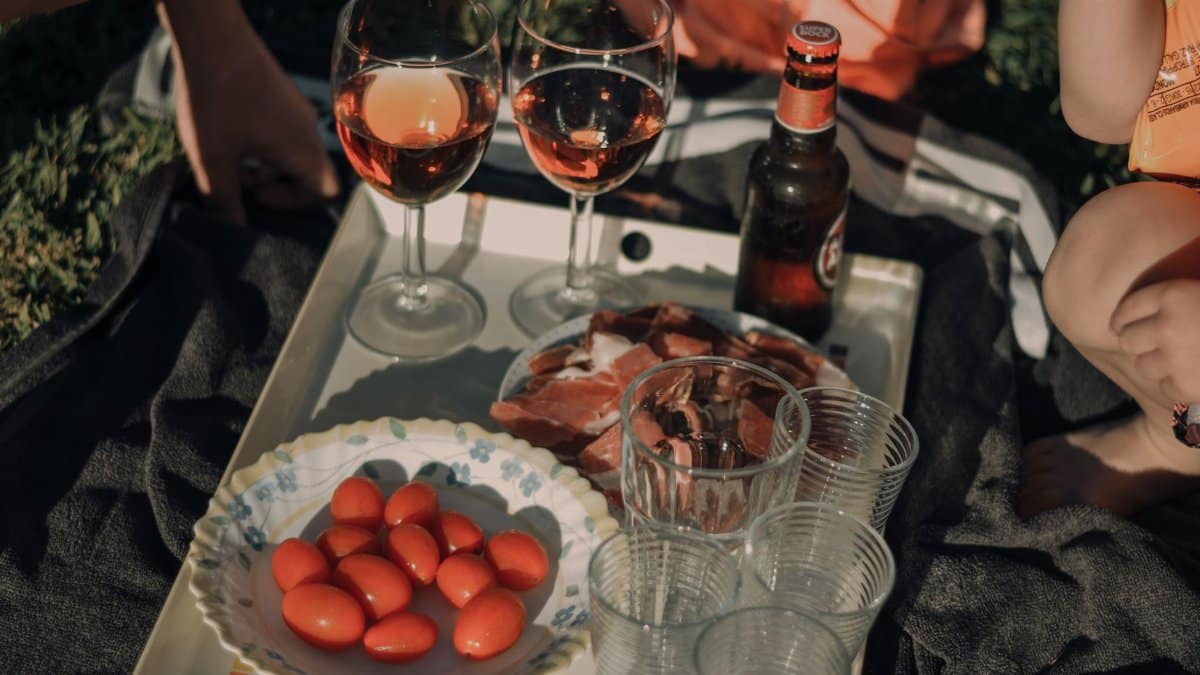 A beautifully arranged outdoor picnic with wine glasses, tomatoes, cold cuts, and a beer bottle on a tray.