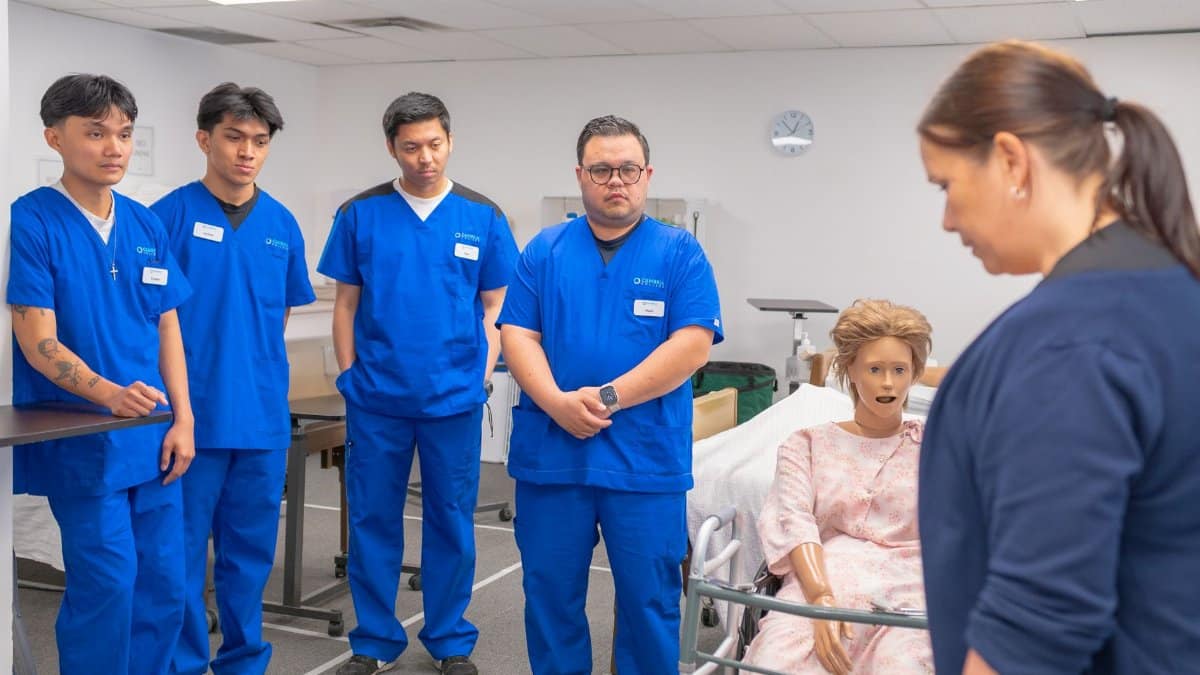 Nursing students in blue scrubs practicing patient care using a medical mannequin.