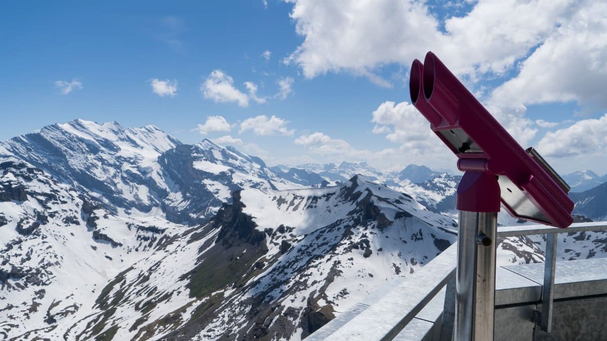 A panoramic view of snow-covered mountain peaks with a telescope on a viewing platform under a bright blue sky.
