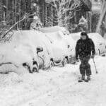 A skier navigates a snowy Boston street, flanked by snow-covered cars and trees, during a heavy snowfall.