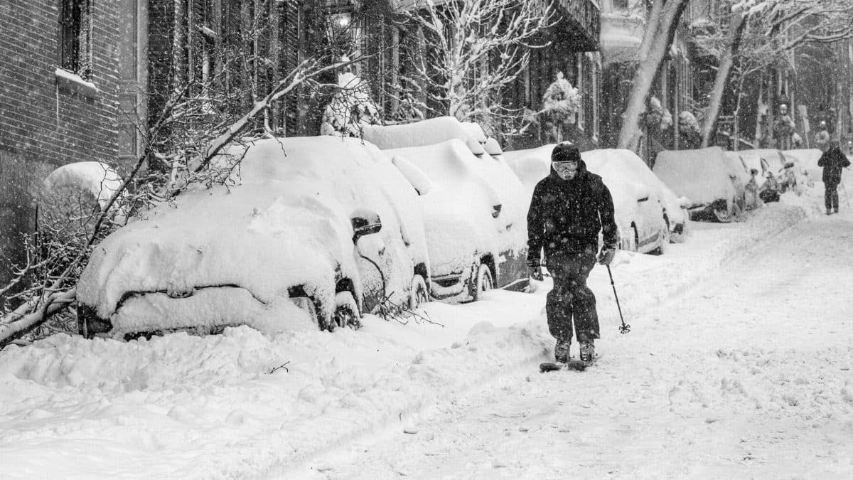 A skier navigates a snowy Boston street, flanked by snow-covered cars and trees, during a heavy snowfall.