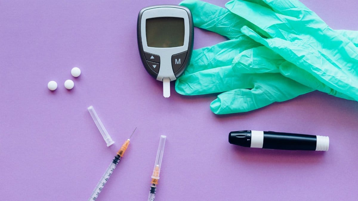 Flat lay of diabetes care tools including glucose meter, syringes, lancet pen, pills, and gloves on purple surface.
