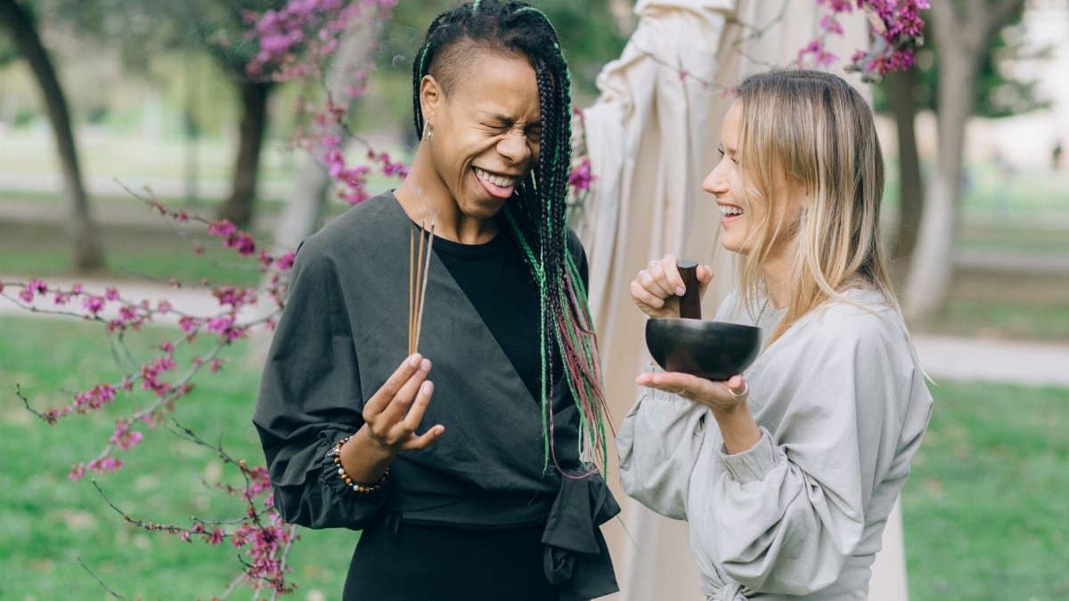 Two women share smiles during a wellness ritual outdoors with a singing bowl.