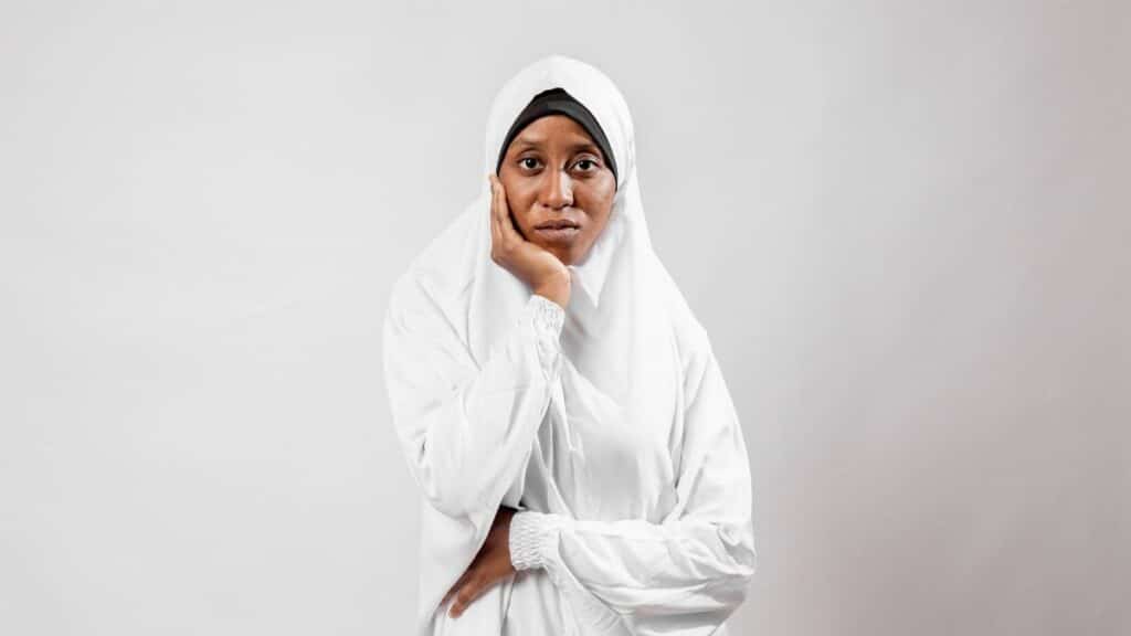 A portrait of a thoughtful woman wearing a traditional white hijab against a plain background.