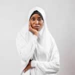 A portrait of a thoughtful woman wearing a traditional white hijab against a plain background.