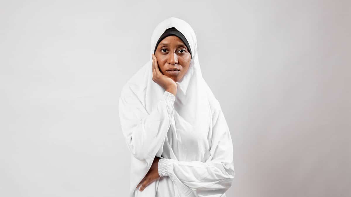 A portrait of a thoughtful woman wearing a traditional white hijab against a plain background.