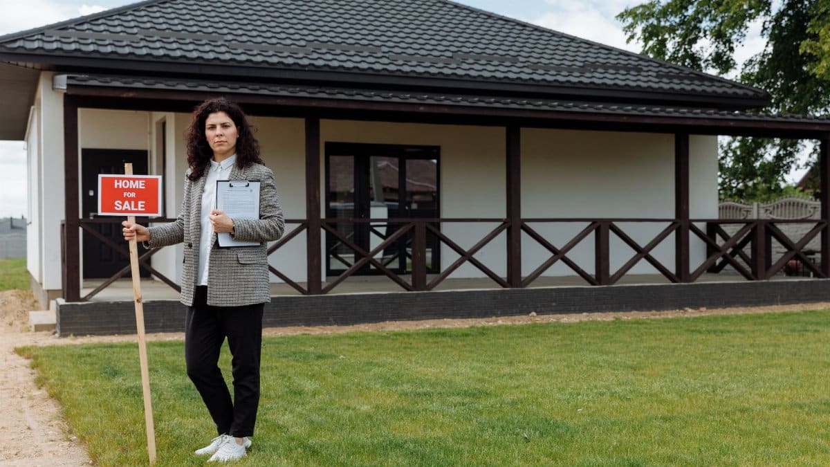 Real estate agent standing outside a house with a 'Home for Sale' sign on a grassy lawn.