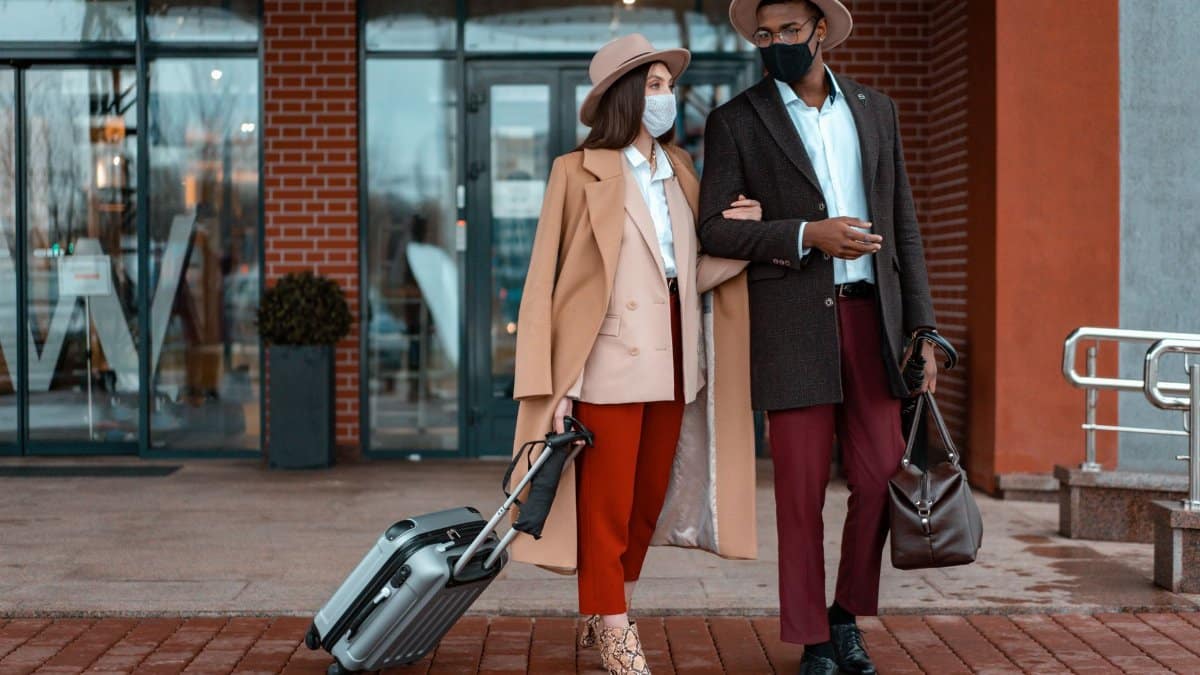 Fashionable couple with face masks at hotel entrance, showcasing travel during pandemic.