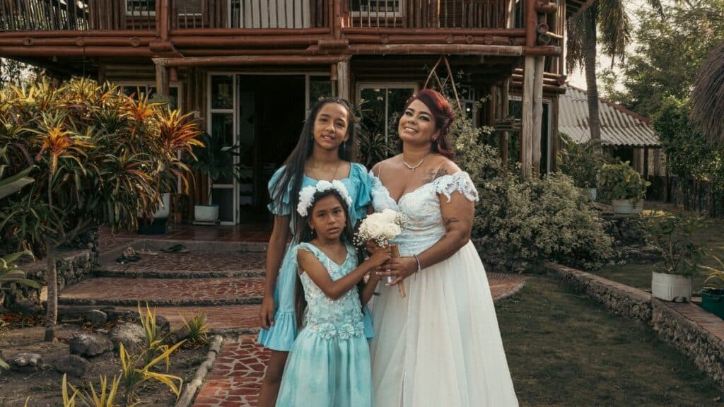 A family of three in elegant dresses, posing outdoors in front of a rustic house.