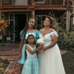 A family of three in elegant dresses, posing outdoors in front of a rustic house.