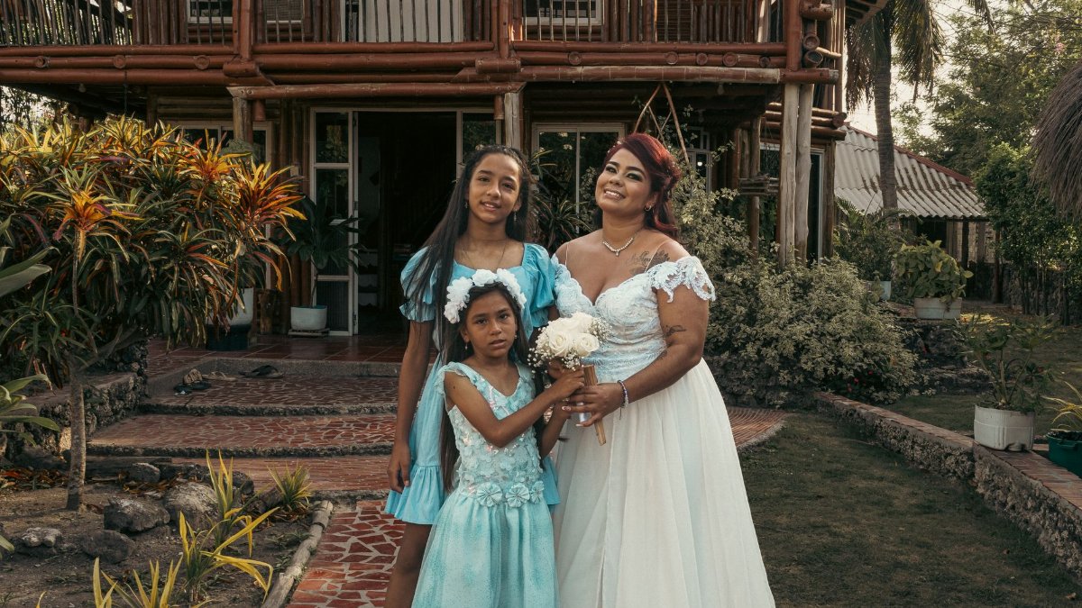 A family of three in elegant dresses, posing outdoors in front of a rustic house.