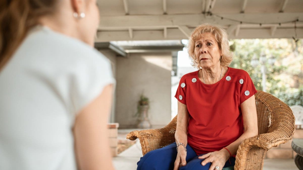 Senior woman in red shirt conversing indoors with younger woman. Daytime scene.
