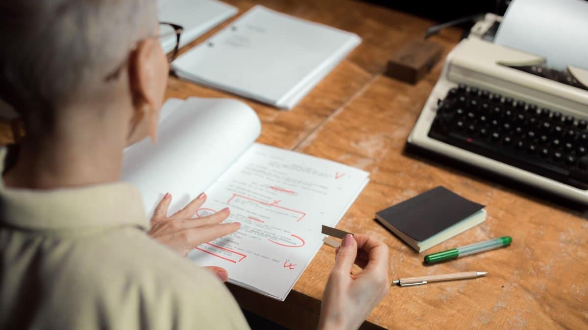 An adult woman reviewing a script with red pen marks at a wooden desk with a typewriter.
