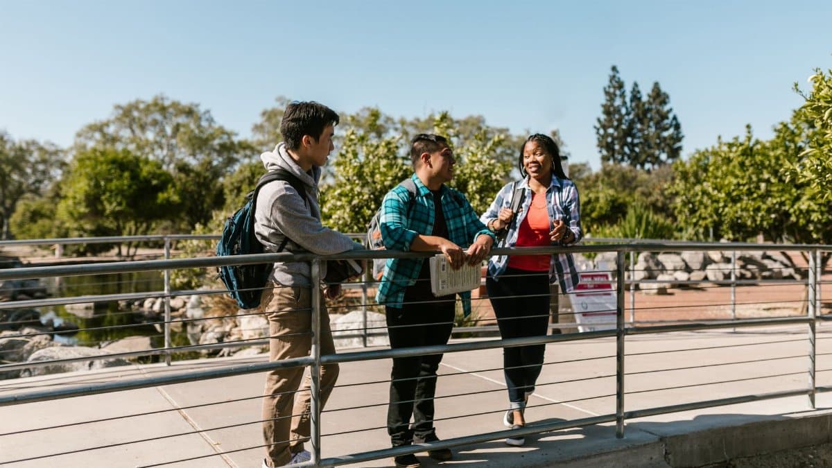 Three college students enjoying a conversation on a sunny day on campus bridge.