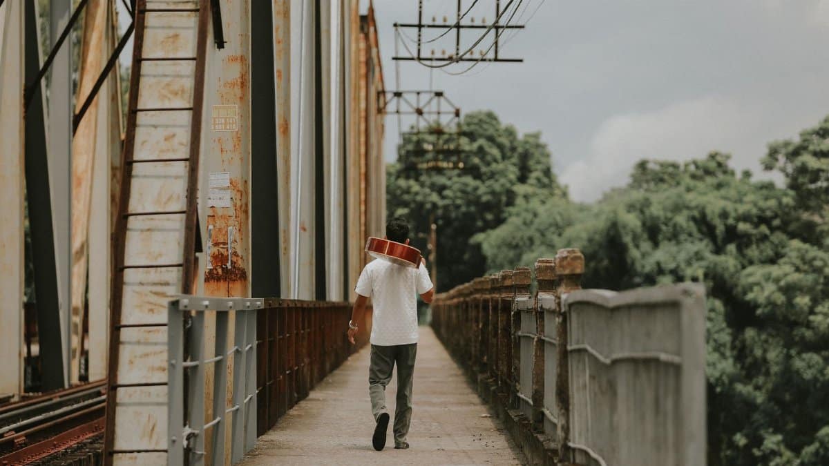 A man walks on a rusty bridge carrying a guitar, symbolizing travel and music outdoors.
