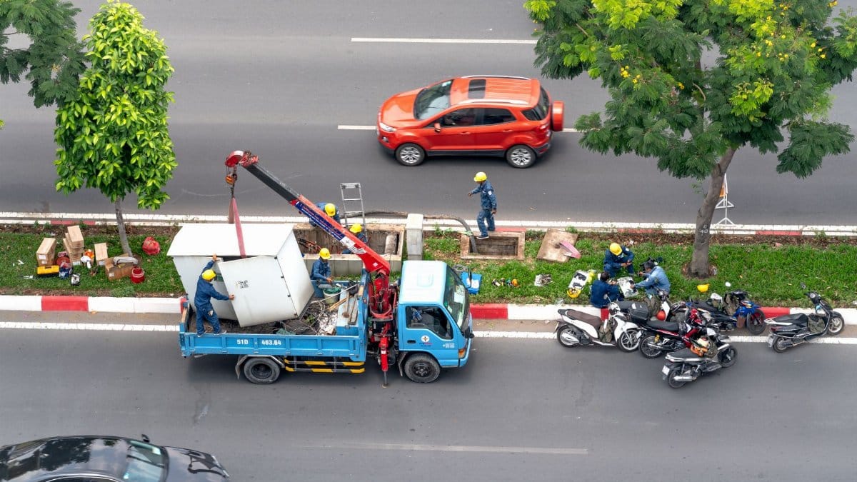 Workers perform maintenance on a busy road using a truck and crane, surrounded by motorcycles.
