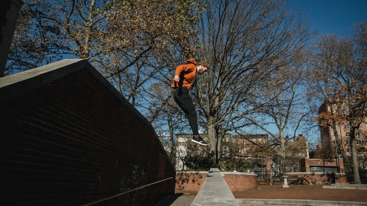 Side view full body sporty male tracer jumping from brick fence in autumn park in modern city