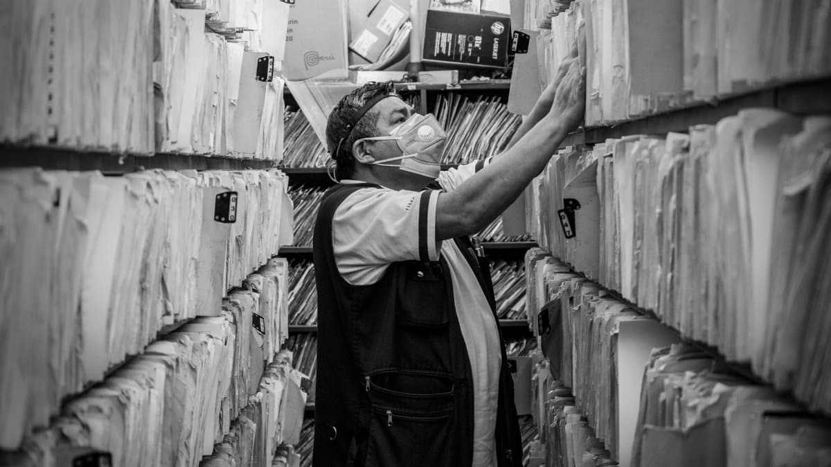 Elderly man sorts through files in an office aisle wearing a face mask.