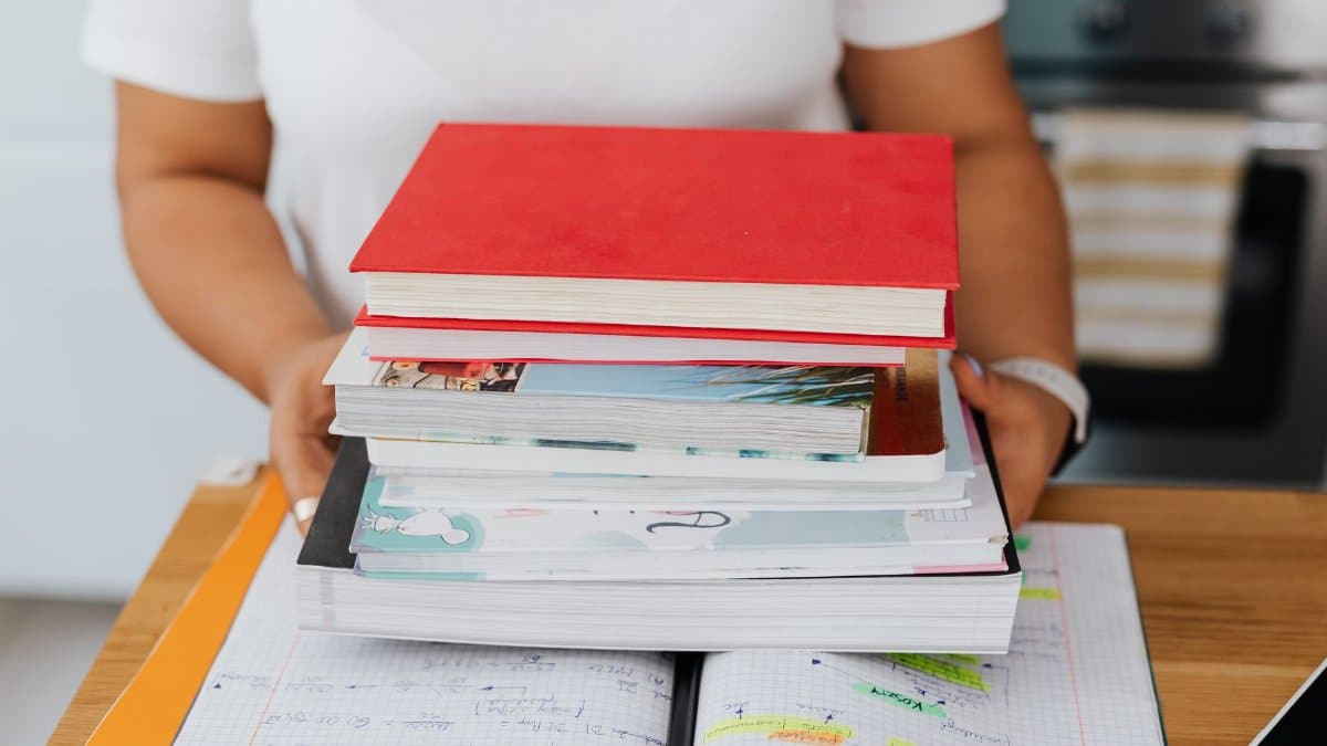 Close-up of a person holding a stack of colorful books over an open notebook.