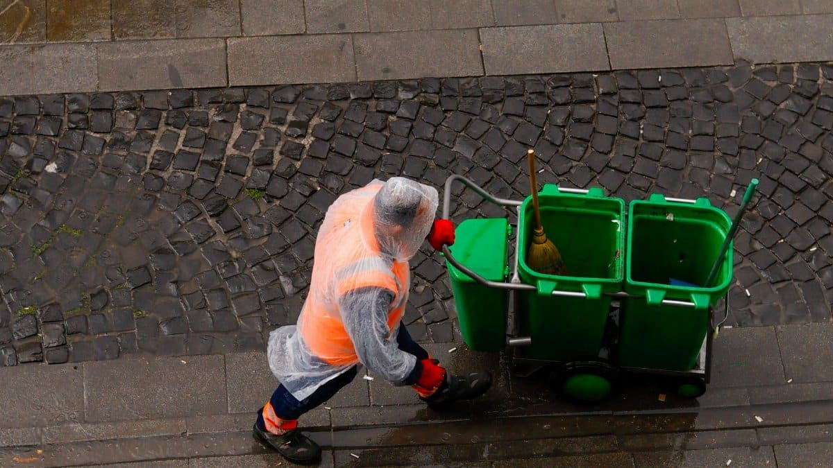A street cleaner in a raincoat pushing a green garbage cart on wet pavement.
