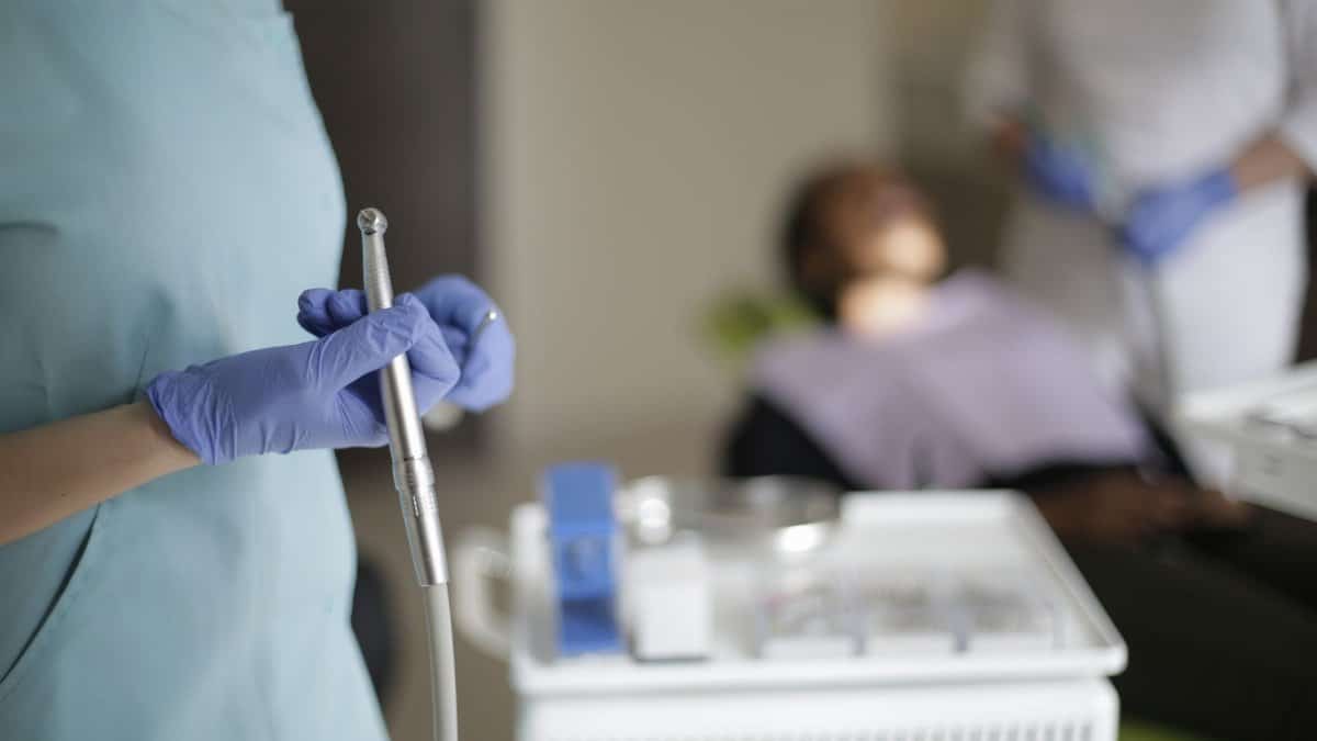 Unrecognizable dentist in uniform and gloves standing with dental drill in selective focus while blurred assistant preparing patient for treating teeth