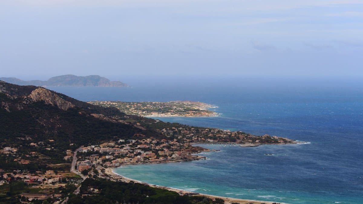 Drone view of coastal city located near hill on grassy coast near rippling sea against blue sky in tropical resort