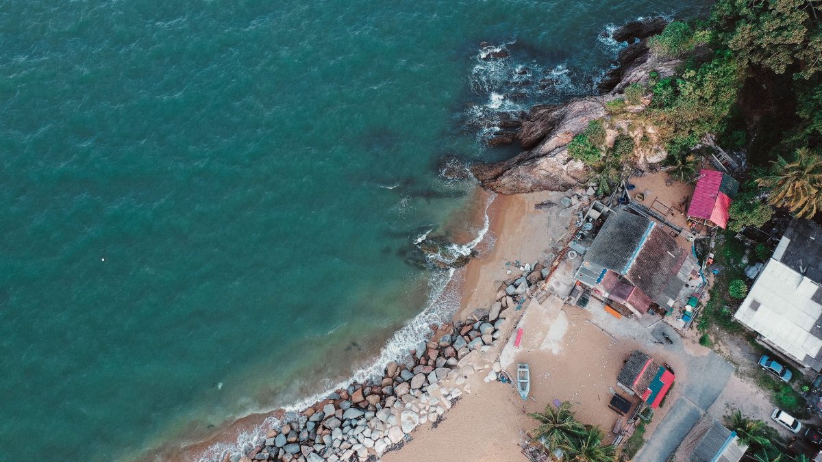 Drone view of a scenic coastal village with vibrant ocean waves meeting the rocky shoreline and lush green trees.