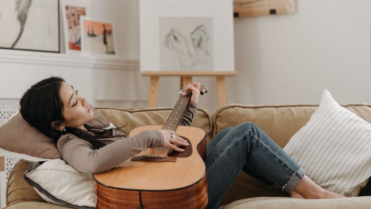 An Asian woman lying on a sofa playing an acoustic guitar in a cozy living room setting.