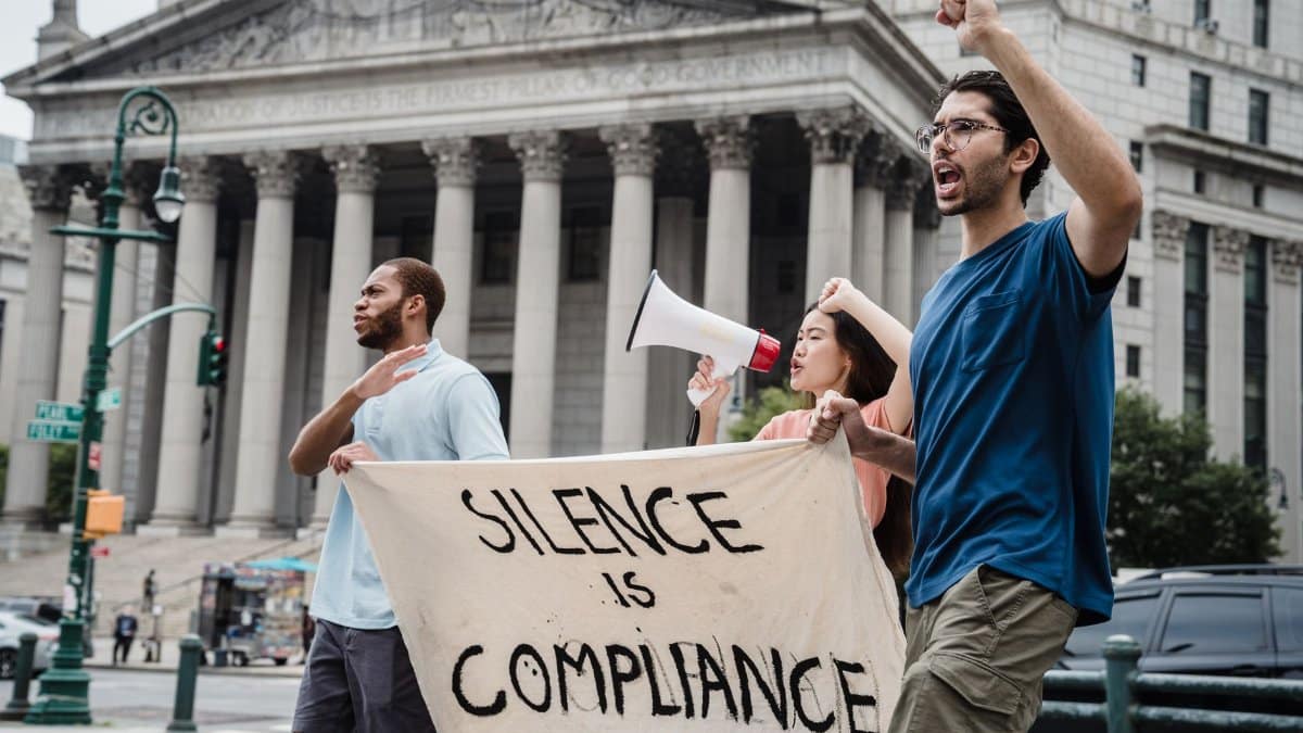 Demonstrators marching with a banner saying 'Silence is Compliance' and a megaphone in front of a courthouse.