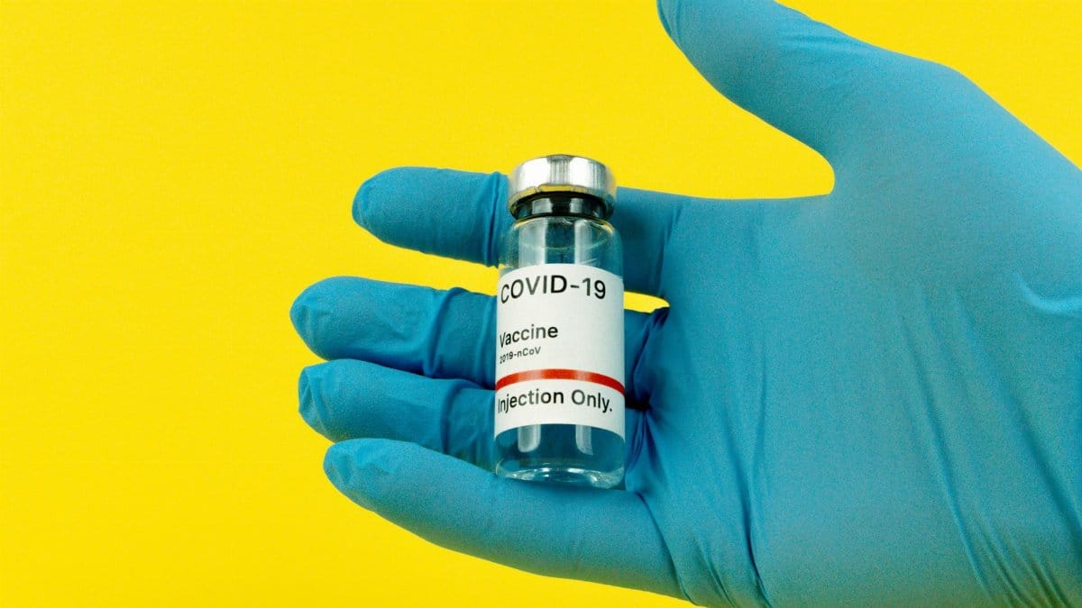 A gloved hand holds a COVID-19 vaccine vial against a bright yellow background.