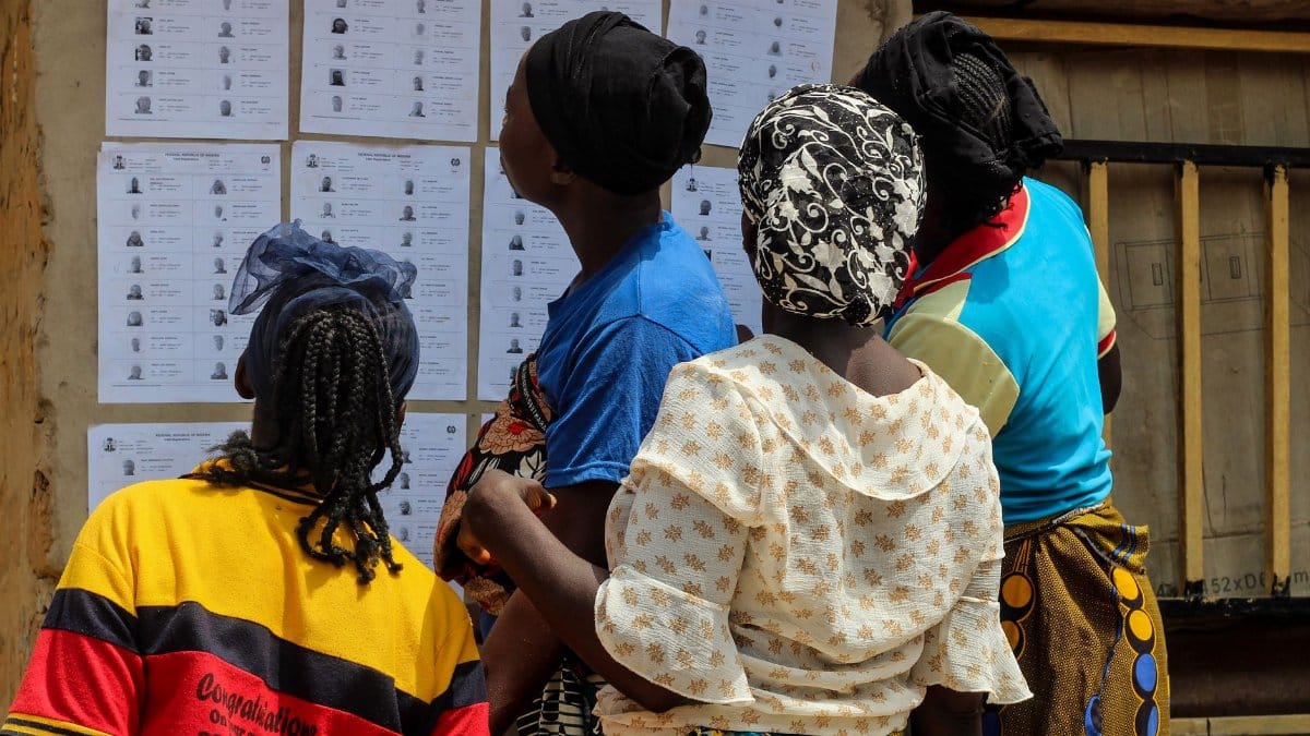 A group of women reading election lists posted on a wall outdoors during the day.