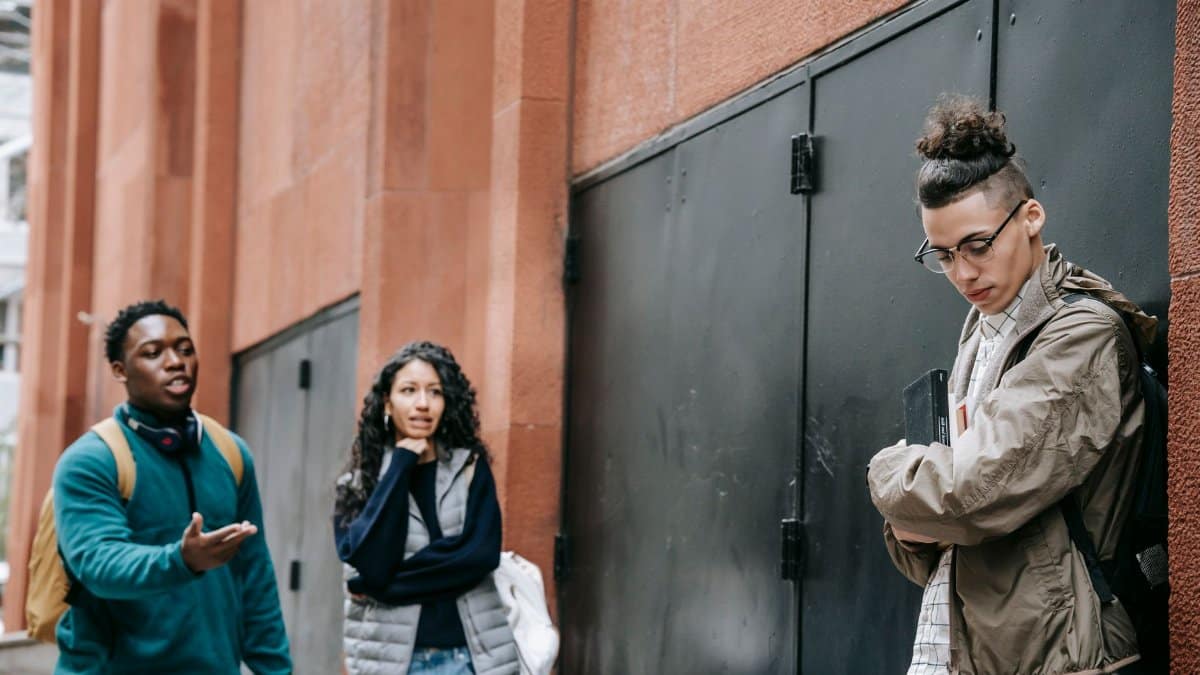 Low angle of aggressive young diverse groupmates bullying shy upset male leaning on building and looking down