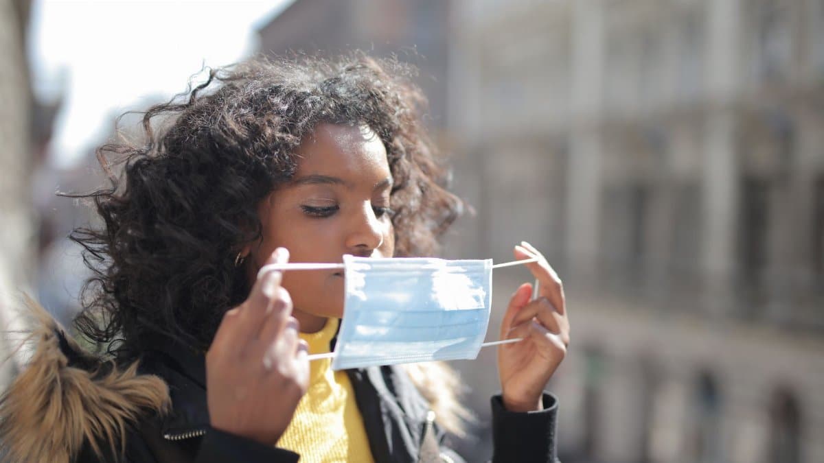 Serious young ethnic lady in warm coat and casual clothes standing on street and putting on medical mask during COVID 19 outbreak
