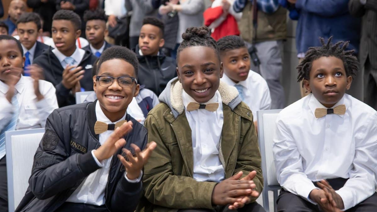 A group of boys dressed smartly, smiling and clapping at an indoor event.