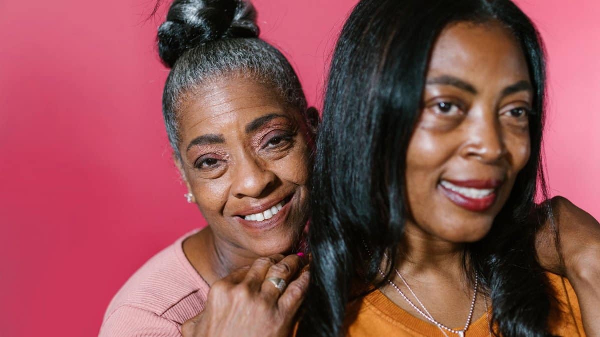 Close-up of two smiling African American women, a senior and an adult, against a pink backdrop.