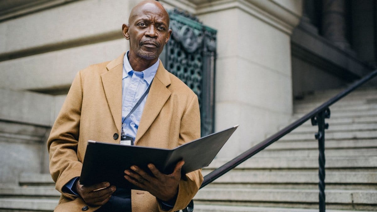 Pensive mature African American male office worker with open folder sitting on staircase handrail while looking away on city street