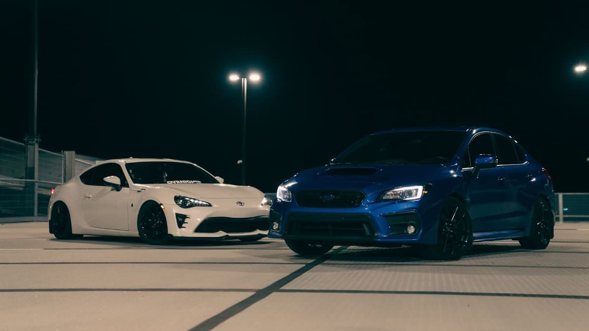 Two sports cars, white and blue, parked on a rooftop at night under streetlights.