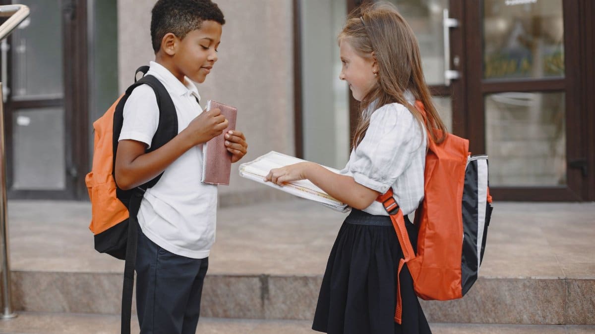 Two children with backpacks exchanging books outside a school building.