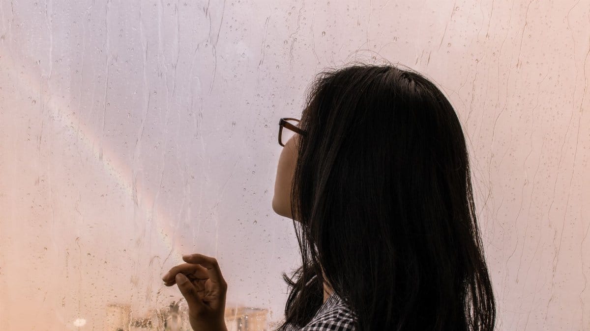 A woman gazes out a rain-speckled window at a faint rainbow, capturing a serene moment of reflection.
