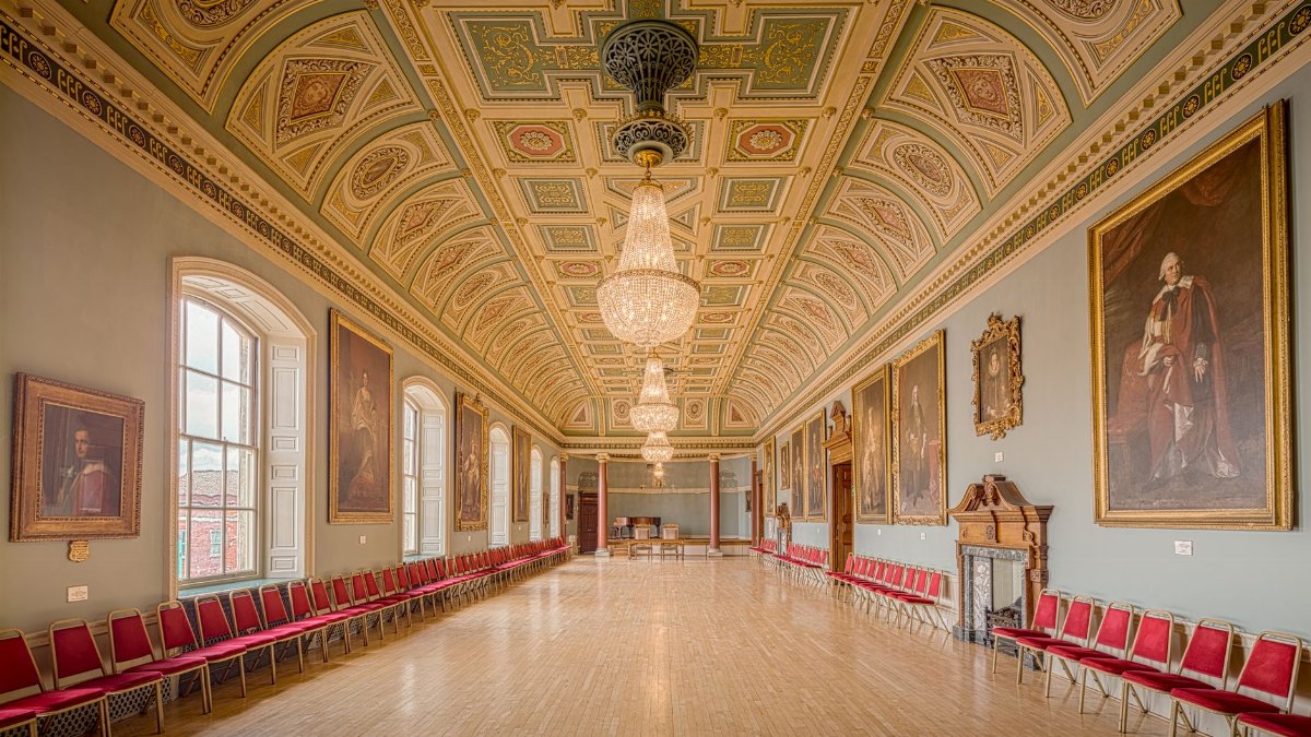 A beautifully preserved historical guildhall interior with ornate chandeliers and large portraits, located in England.
