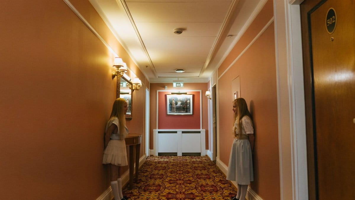 Two blonde girls standing in a vintage hotel corridor, reflecting symmetry and nostalgia.