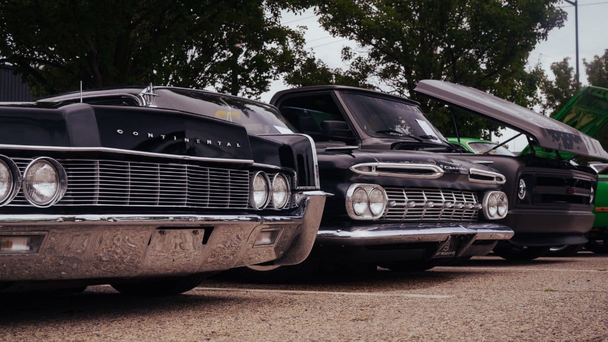 A lineup of classic cars displayed outdoors under trees, showcasing vintage designs.