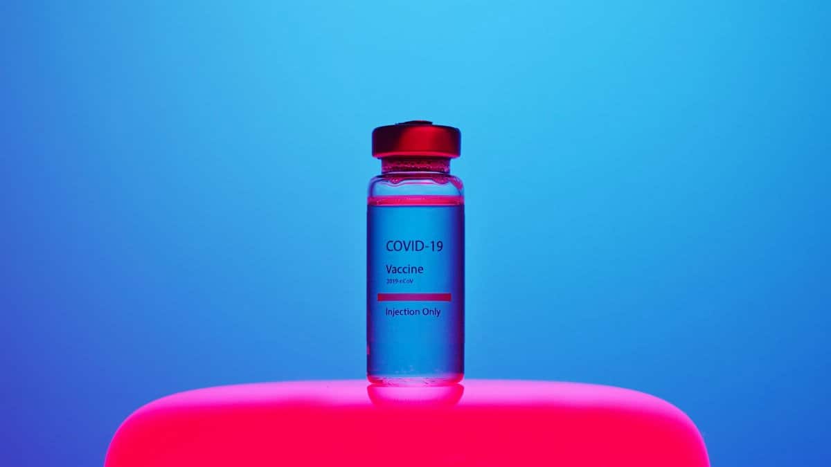 A COVID-19 vaccine vial sits on a bright, blue and pink background. Modern and impactful.